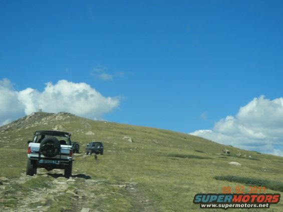White 1996 Ford Bronco rear view on a grassy mountain road with another SUV behind