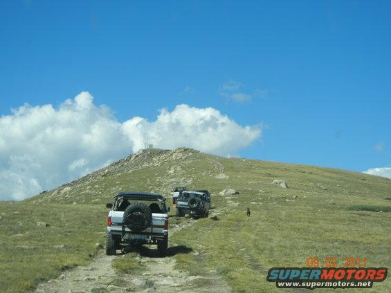 1996 Ford Bronco and two other SUVs driving up a rocky mountain path under a blue sky with clouds