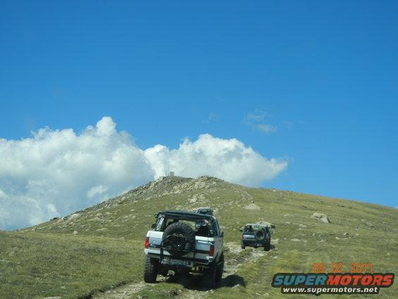 White SUV with spare tire on rear, driving on dirt road up grassy hill with another vehicle behind