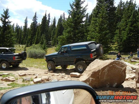 Dark green 1996 Ford Bronco climbing over a large rock on a dirt trail with trees in the background