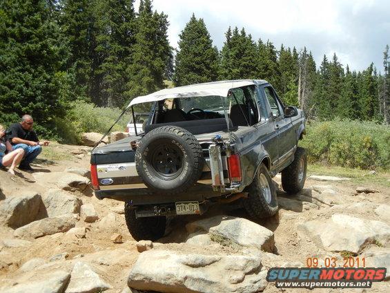 Rear view of a dark gray Ford Bronco with a white soft top on a rocky trail