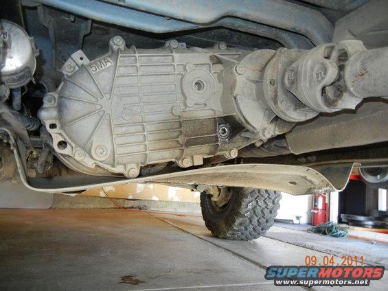 Underside view of a vehicle's transfer case and driveshaft. A tire is visible