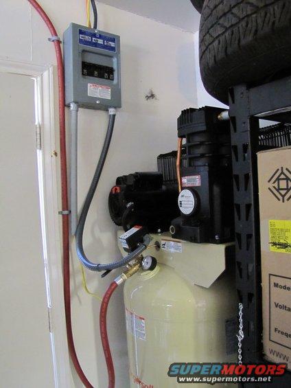 Air compressor with gauge and electrical panel on wall. Tire on shelf above