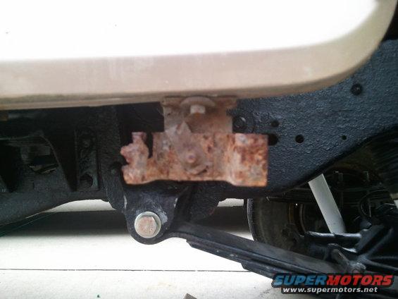 Close-up of a rusted bracket attached to the frame of a vehicle, with part of the body and suspension visible