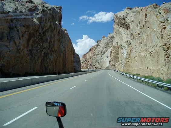 Truck side mirror reflecting an orange object, with a highway passing through a rocky canyon under a blue sky