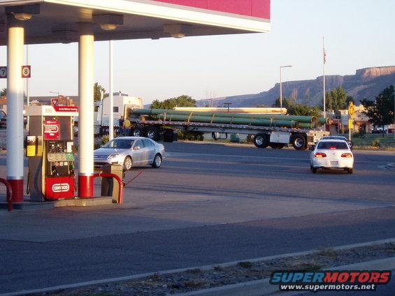 Silver sedan parked at gas station, semi-truck with pipes on trailer, white car driving away