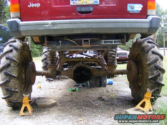 Rear view of a red Jeep SUV lifted with massive tires, jack stands under the rear axle. Another vehicle is visible in the background