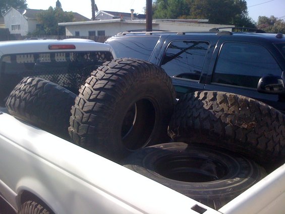 Pickup truck bed loaded with off-road tires. Dark SUV parked behind