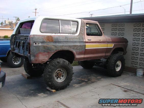Lifted 1978 Ford Bronco with large off-road tires, showing rust and missing rear body panels. Another blue truck is partially visible to the left