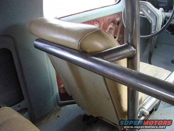 Roll cage tubing crosses in front of tan vinyl seats inside a vehicle. The steering wheel and dashboard are partially visible