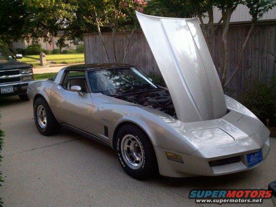Silver 1982 Chevrolet Corvette with hood open, parked on a driveway next to a black pickup truck