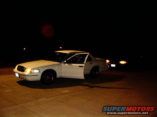 White sedan with driver's door open, parked at night. Headlights on, dark background with lights