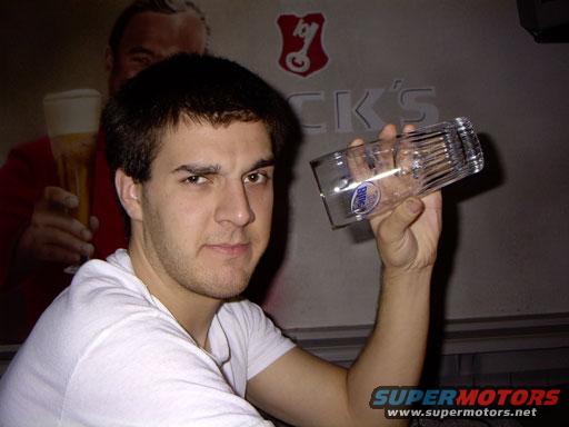 Man holds a clear beer glass with a blue label. Background shows a poster of a man holding a beer