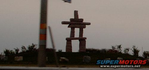 Inukshuk statue on a grassy hill with a utility pole in the foreground. Supermotors.net logo in the corner