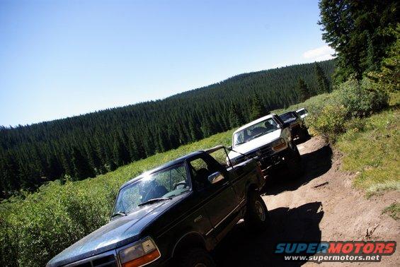 Dark green 1996 Ford Bronco on a dirt road with two other trucks behind it, heading into a forest