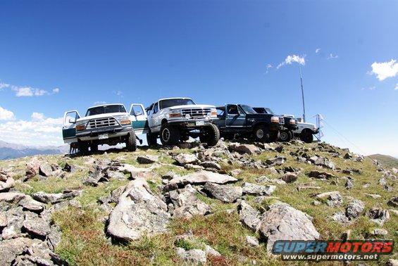 Four SUVs parked on a rocky hilltop under a blue sky. The lead vehicle is a white 1996 Ford Bronco