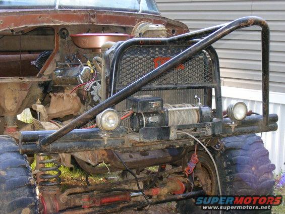 Front of a heavily modified vehicle with a winch, large tires, and a custom bumper. Engine components are visible