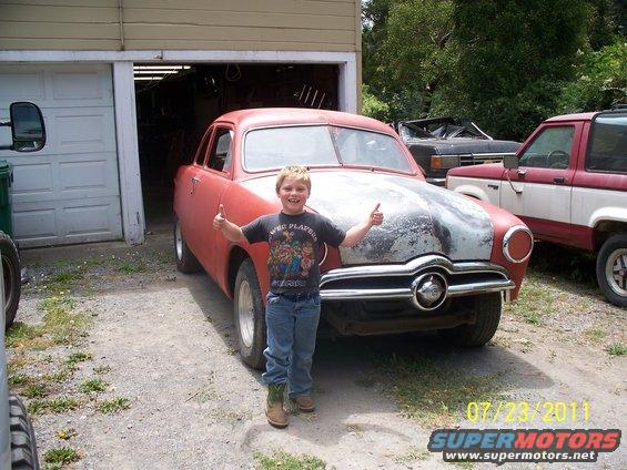 Red coupe with a boy giving thumbs up in front of a garage