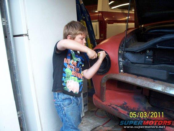Boy in jeans and t-shirt works on a red vehicle's headlight. Another red vehicle is visible in the background
