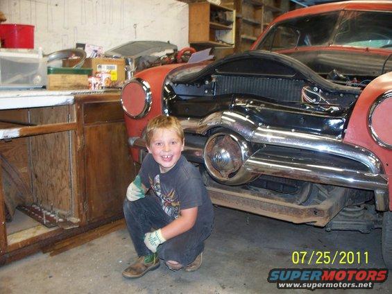 Boy wearing work gloves crouches in front of a vintage car's front end, which is partially disassembled in a garage
