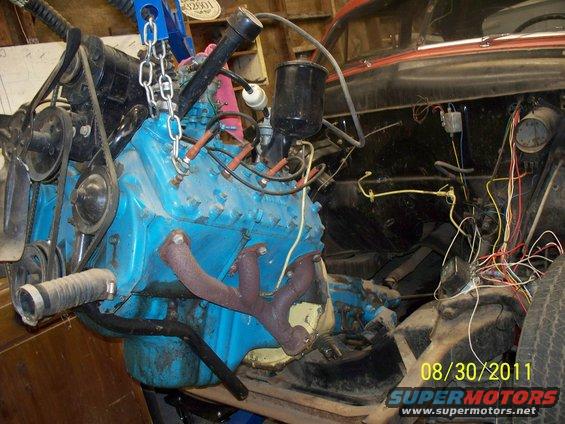 Blue engine block with exhaust manifold, carburetor, and wiring harness. Engine is suspended by a chain hoist