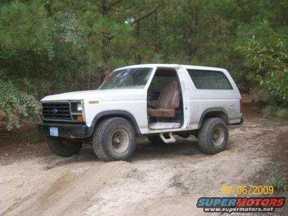 White Ford Bronco with driver's door open, parked on a dirt path with trees in the background