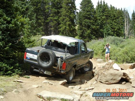 1996 Ford Bronco navigating a rocky mountain trail with a woman taking photos in the background