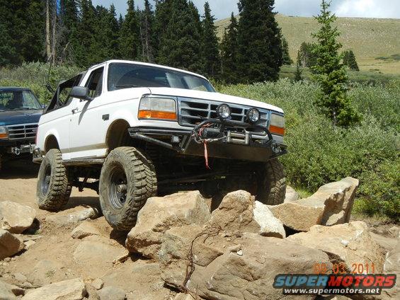 White 1996 Ford Bronco with aftermarket bumper and winch navigating rocky terrain. Another vehicle is visible behind it