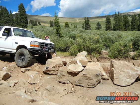 White 1996 Ford Bronco navigating a rocky off-road path with a person leaning on the driver's side door