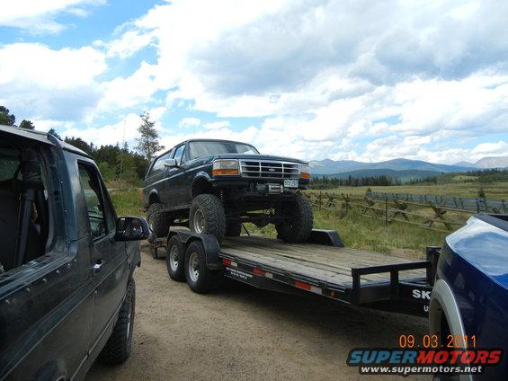Dark green Ford Bronco on a trailer, lifted with large tires. Another vehicle is partially visible to the left