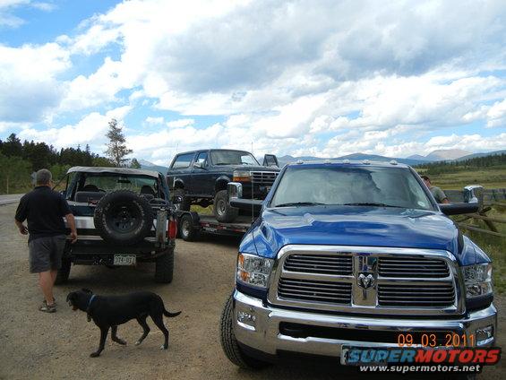 Dark blue Ford Bronco on a trailer, towed by a blue pickup truck. A man and a dog are nearby