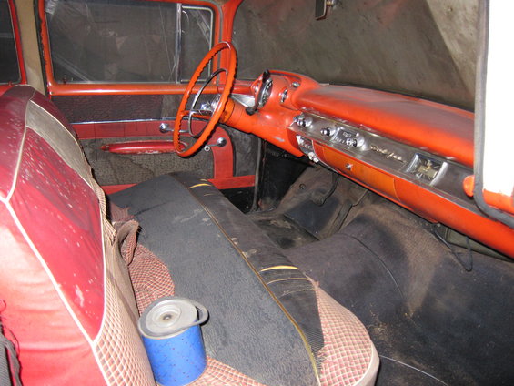 Interior of a vintage car with red and black seats, orange dashboard, and steering wheel. A blue cylindrical object sits on the seat