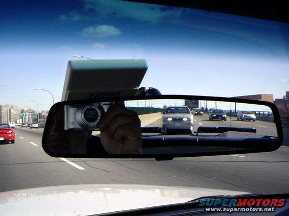Reflection in rearview mirror shows traffic on highway; camera is visible in reflection