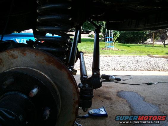 Close-up of vehicle suspension components, including a coil spring, shock absorber, and brake rotor, with a power tool on the ground