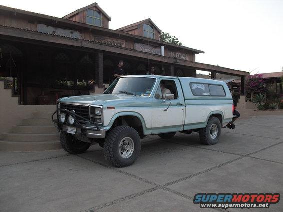Light blue and white Ford F-Series pickup truck with camper shell, bull bar, and off-road tires parked in front of a building
