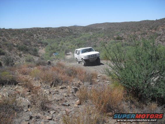 White 1987 Ford Bronco driving off-road, another blue vehicle in distance