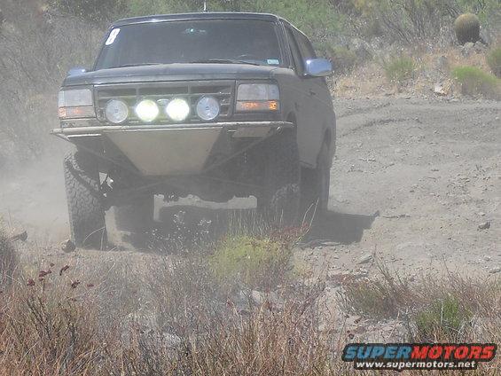 Dark-colored 1992 Ford Bronco with aftermarket bumper and lights driving on a dirt road