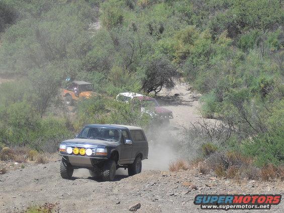 Dark blue Ford Bronco with yellow auxiliary lights on front bumper, driving on a dirt road with other off-road vehicles behind it