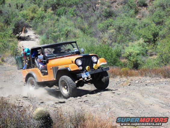 Orange Jeep CJ-7 driving on a dirt road, kicking up dust. License plate visible