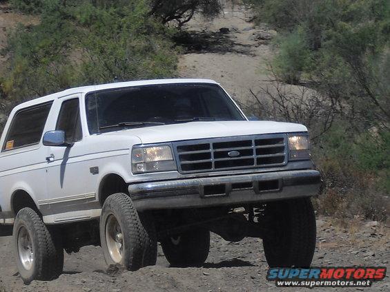 White Ford Bronco driving on a dirt road, front 3/4 view. The vehicle has a chrome grille and bumper
