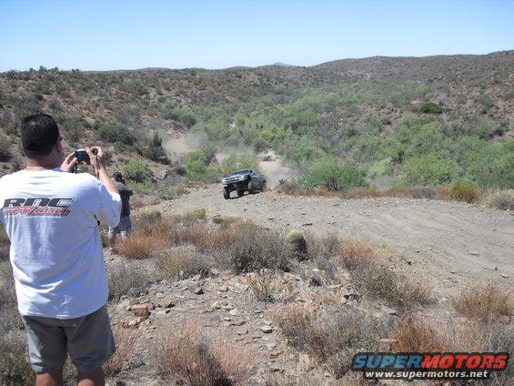 guy taking photo of off-road truck kicking up dust on a dirt path in a dry, hilly area