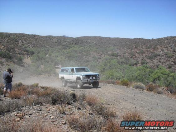 Two-tone blue and white Ford Bronco driving on a dusty dirt road