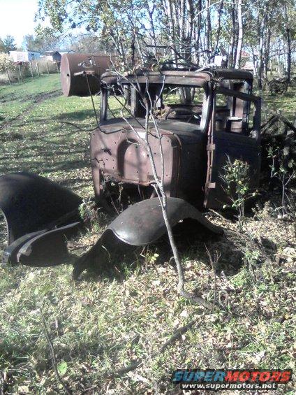 Rusty truck cab and detached fenders in a grassy field with trees in the background