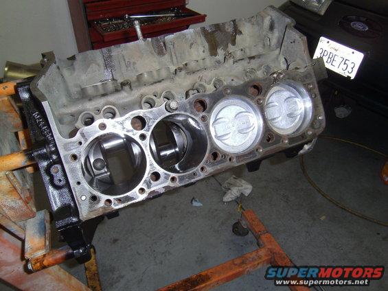 Engine block with three pistons visible, mounted on an orange engine stand. A license plate is partially visible in the background