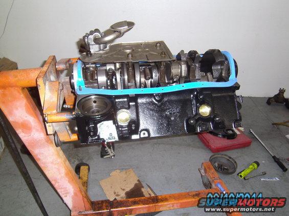Engine block on an orange engine stand, with crankshaft and oil pump visible. Blue gasket installed