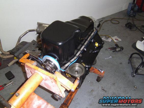 Engine block on an orange stand, with exhaust manifold and oil pan attached. Tools and equipment are visible in the background