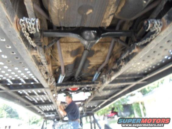 Undercarriage of a vehicle on a car hauler, showing chains, exhaust pipes, and differential