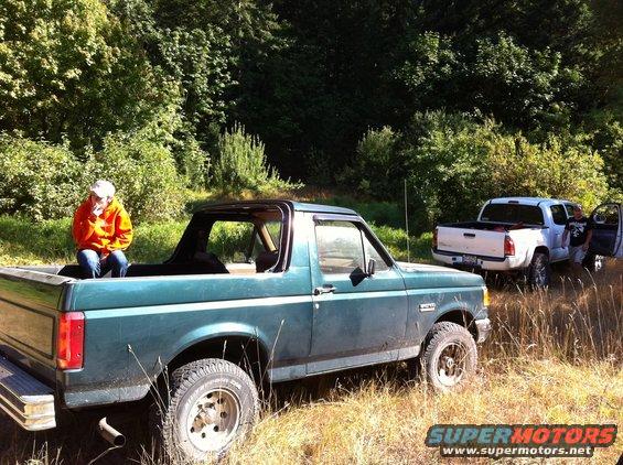 Green Ford Bronco, 1987 model year, with a person in the bed, parked next to a white pickup truck