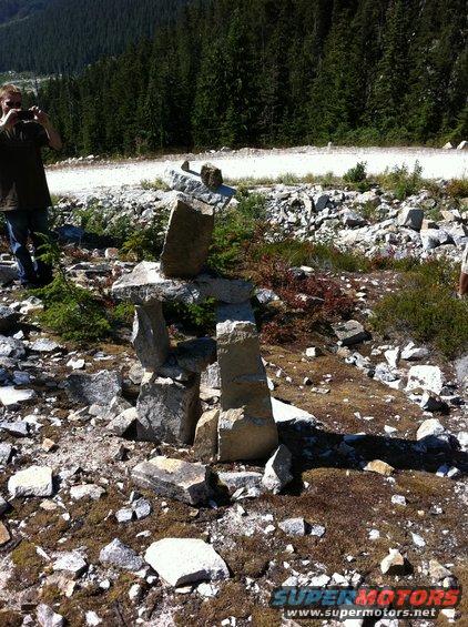 Person taking photo of stone inukshuk on rocky hillside with trees and dirt road in background