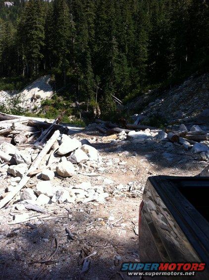 Truck bed edge, dark color, seen from above and behind, parked on a rocky, wooded trail